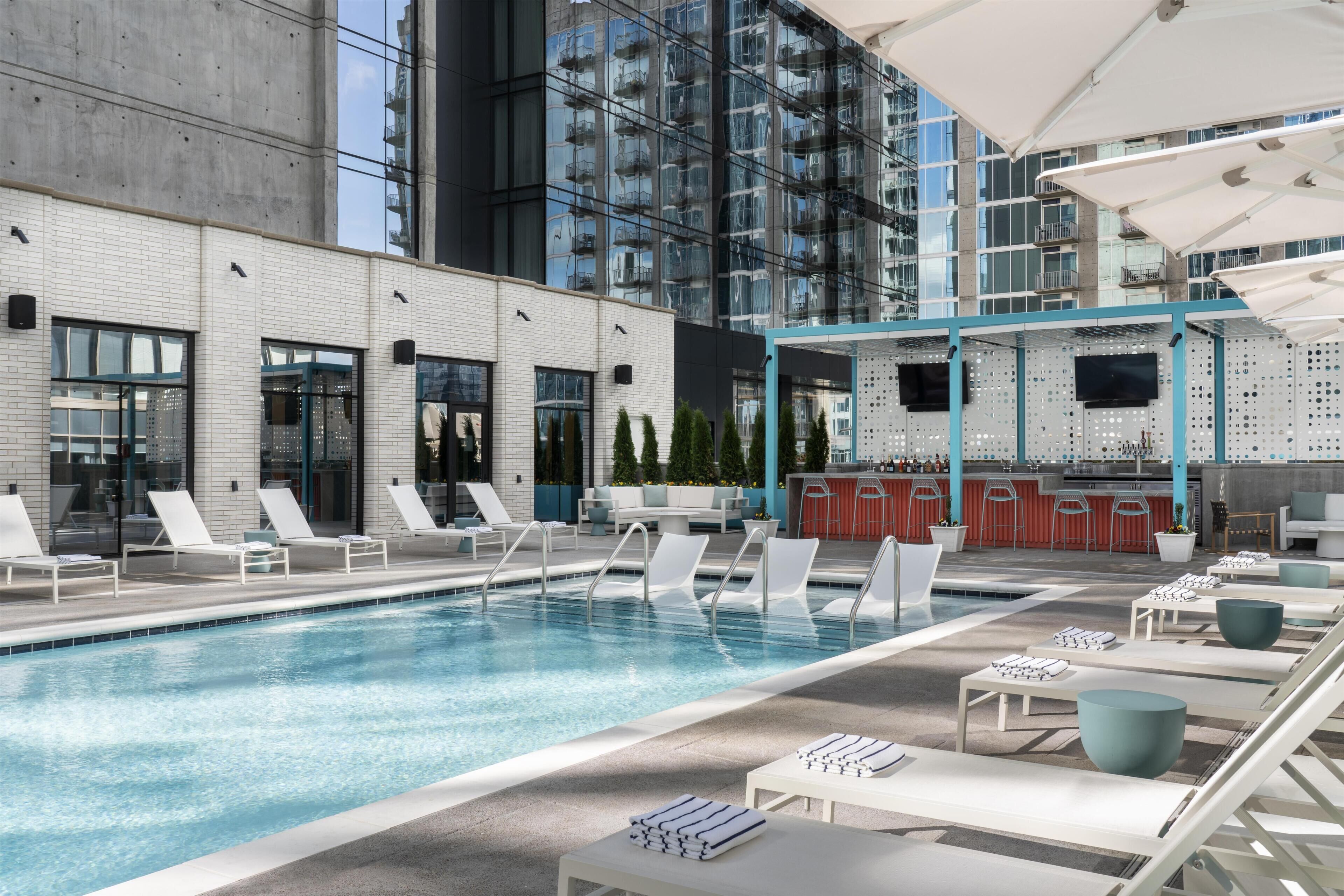 A rooftop pool area with lounge chairs, a bar, and a view of a modern building.