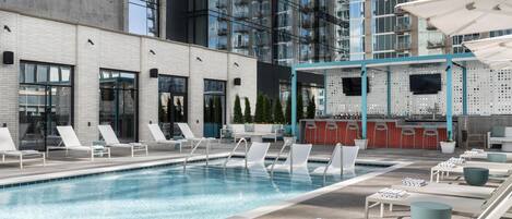 A rooftop pool area with lounge chairs, a bar, and a view of a modern building.