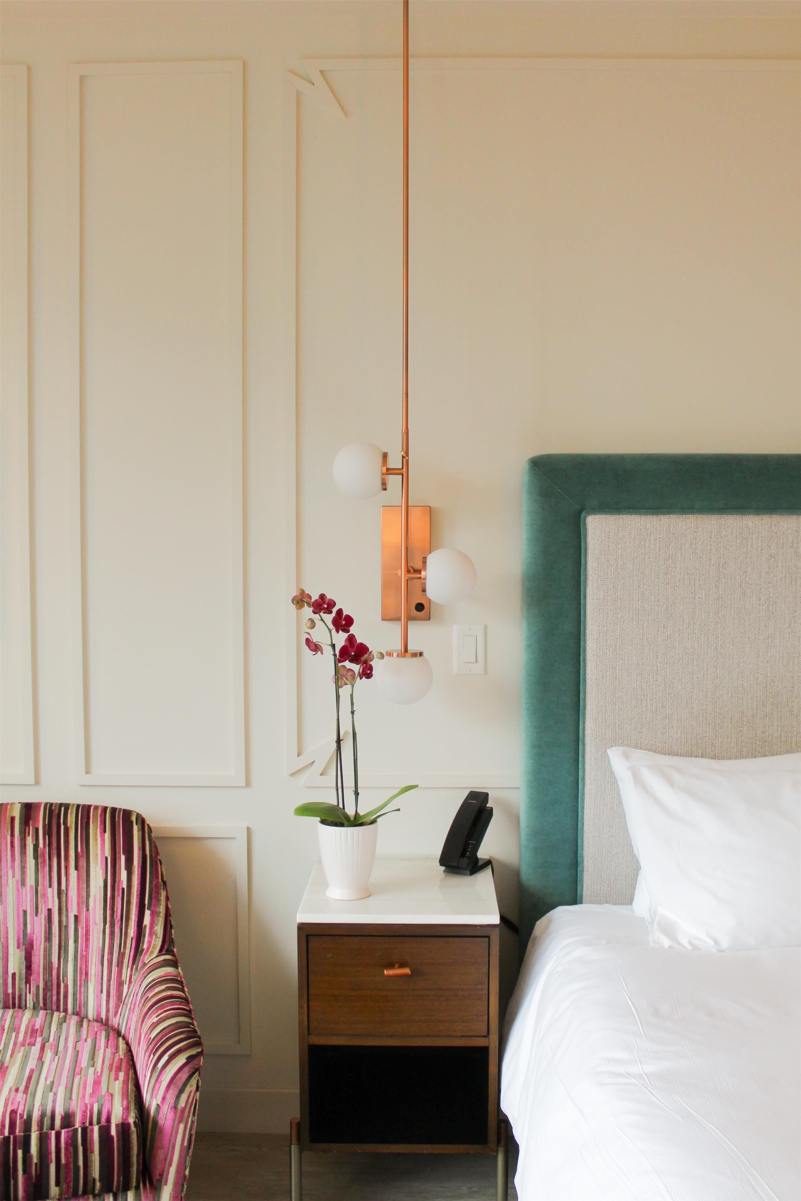 Quarto de hotel com uma cama, mesa de cabeceira com telefone, uma cadeira e luminária na parede.