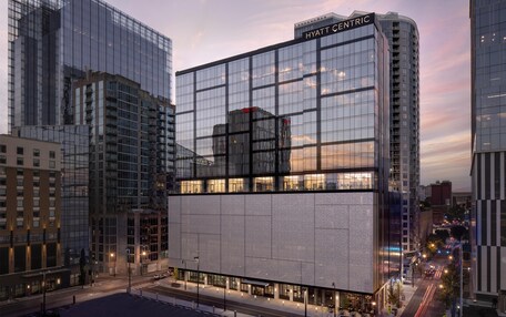 A modern hotel with a glass facade, a prominent 'HYATT CENTRIC' sign, and a street view with parked cars and a clear sky.