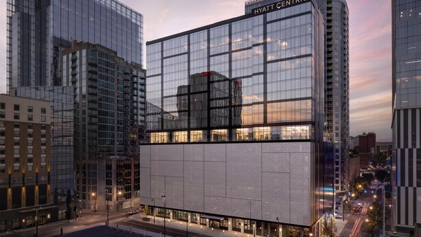 Un hotel moderno con fachada de vidrio, un llamativo letrero de 'HYATT CENTRIC' y vista a la calle con coches estacionados y cielo despejado.