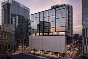 Un hôtel moderne avec une façade en verre, une enseigne « HYATT CENTRIC » bien visible et une vue sur la rue, avec des voitures garées et un ciel dégagé.
