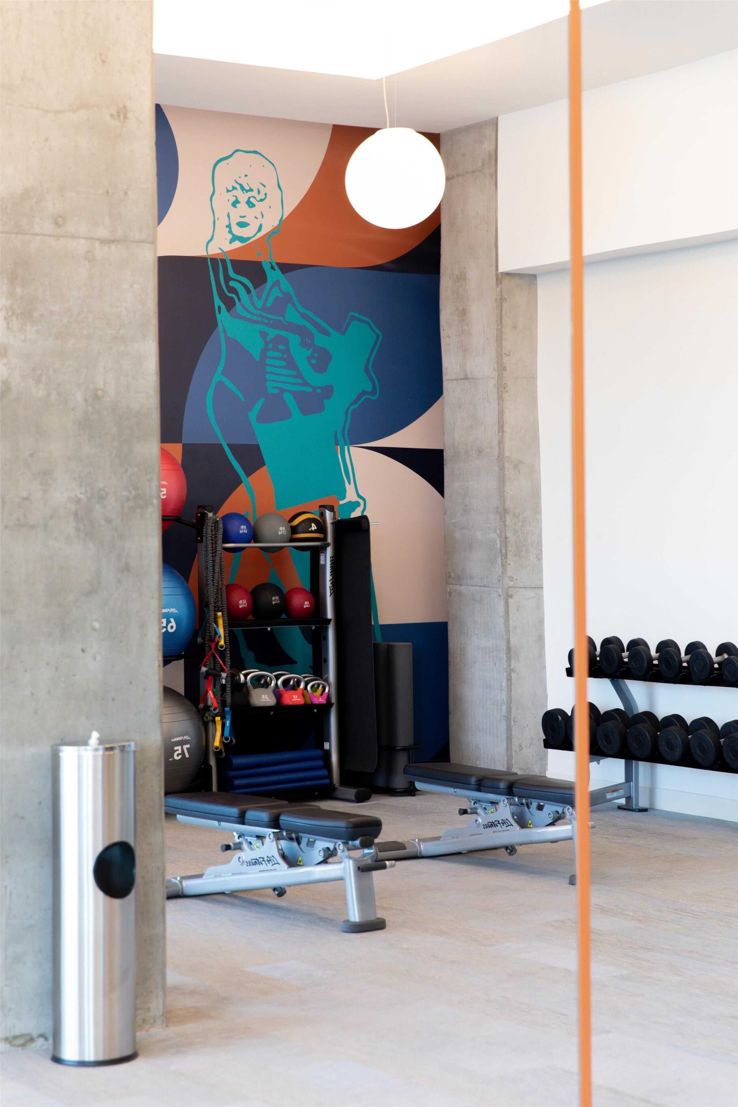 Une salle de sport équipée de matériel d’exercice, un mur orné d’œuvres d’art abstrait et un pilier en béton.