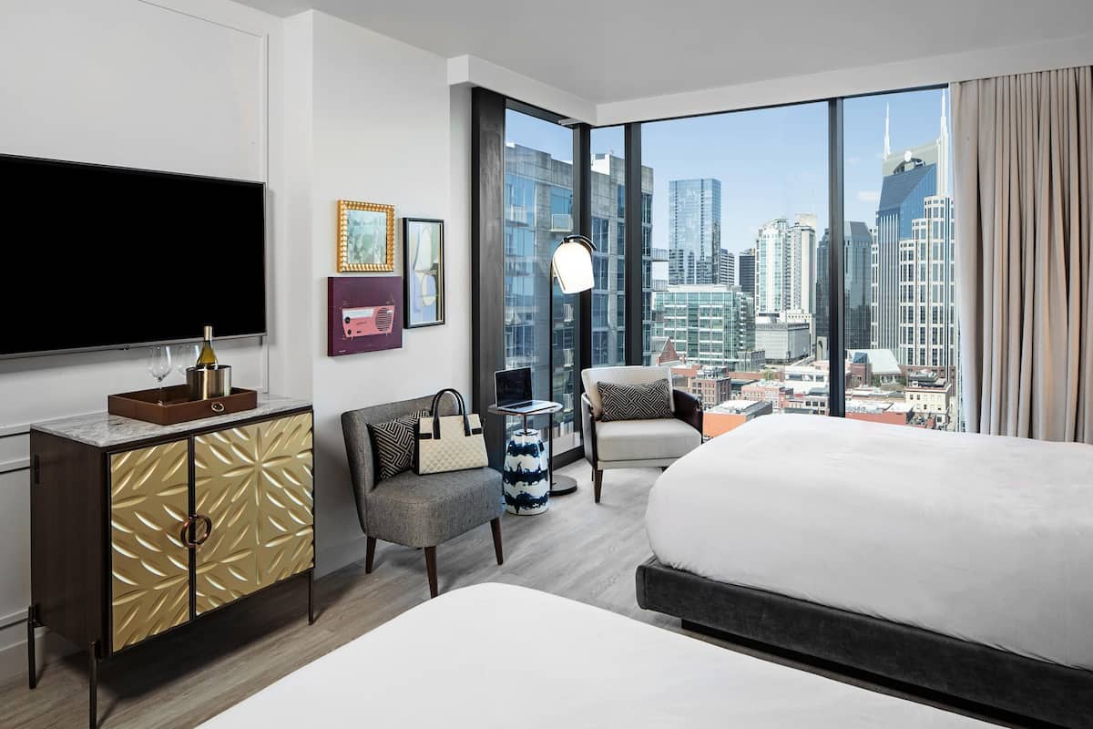 A modern hotel room with a large bed, a TV, a desk with a laptop, a chair, and a city view.