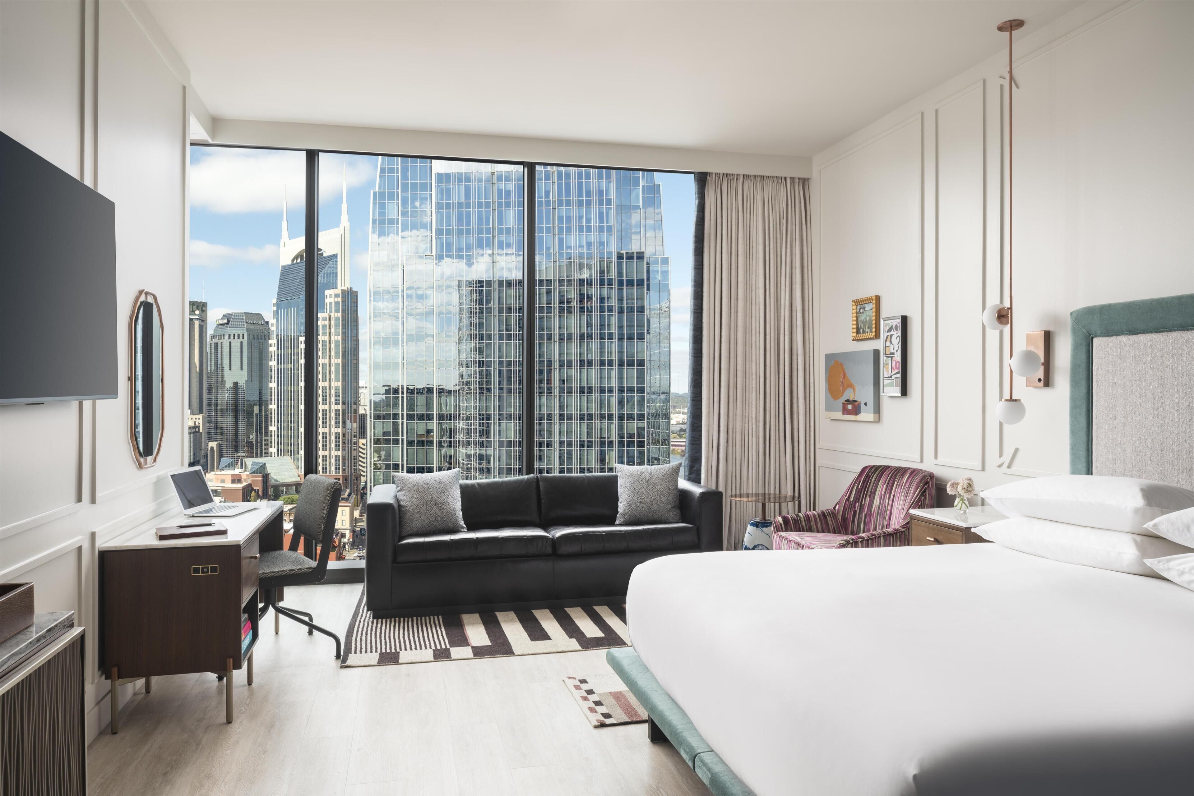 A hotel room with a large bed, a desk with a laptop, a sofa, and a view of city skyscrapers.