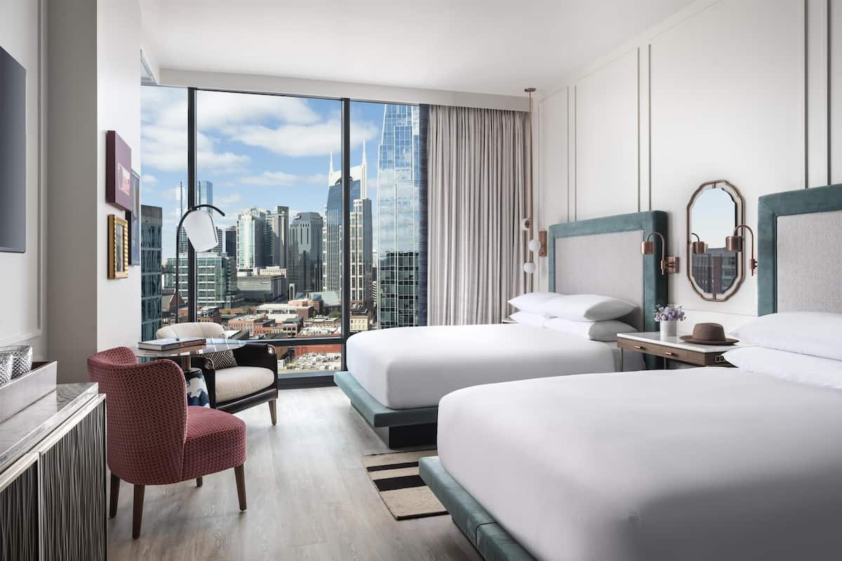A hotel room with two beds, a desk, a chair, and a large window with a city view.