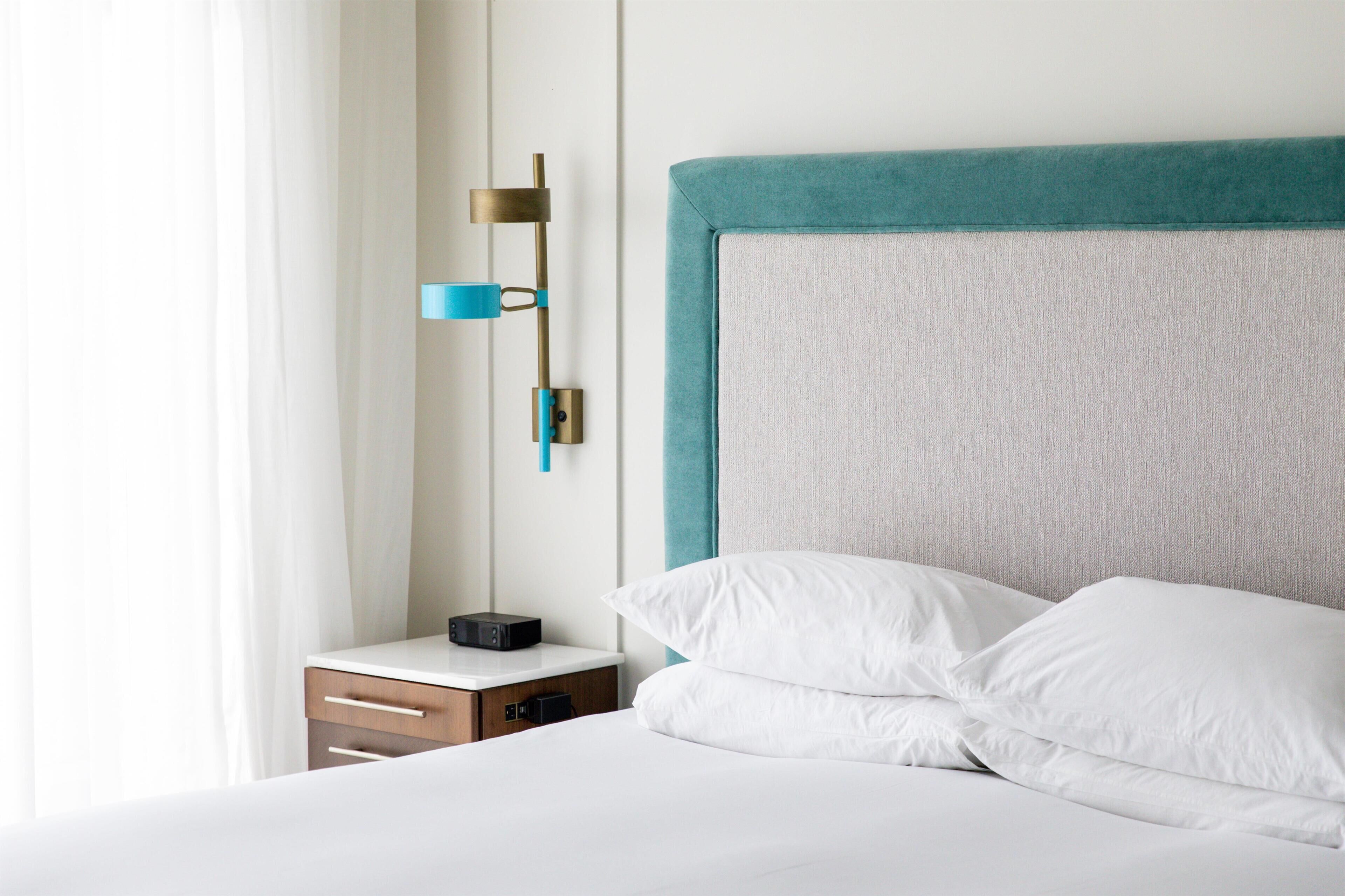 Una cama bien hecha con ropa de cama blanca, una mesita de noche con un pequeño dispositivo negro y una lámpara fijada a la pared.