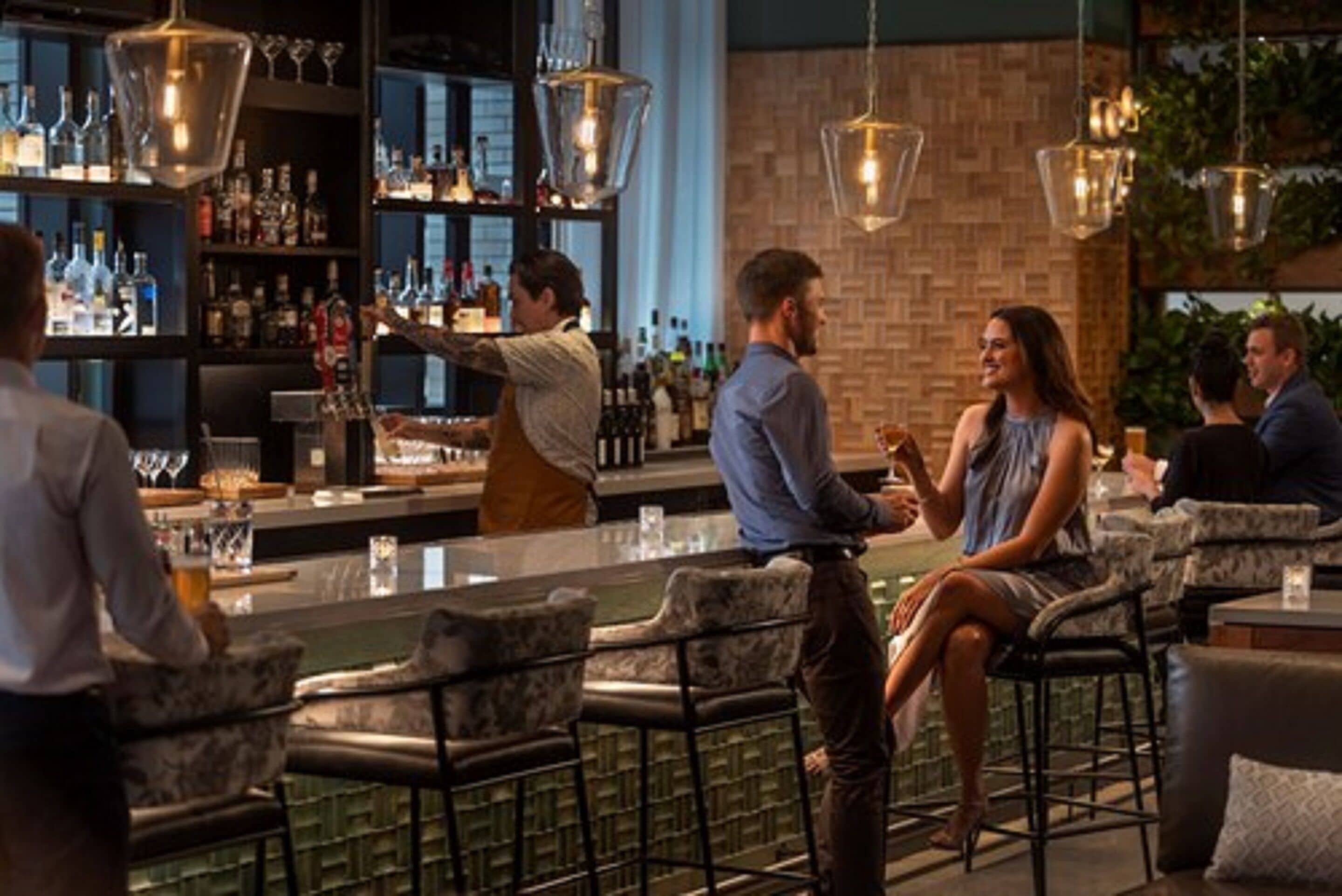 A bar area with a counter, bar stools, and a bartender. There are shelves with bottles and glasses in the background.