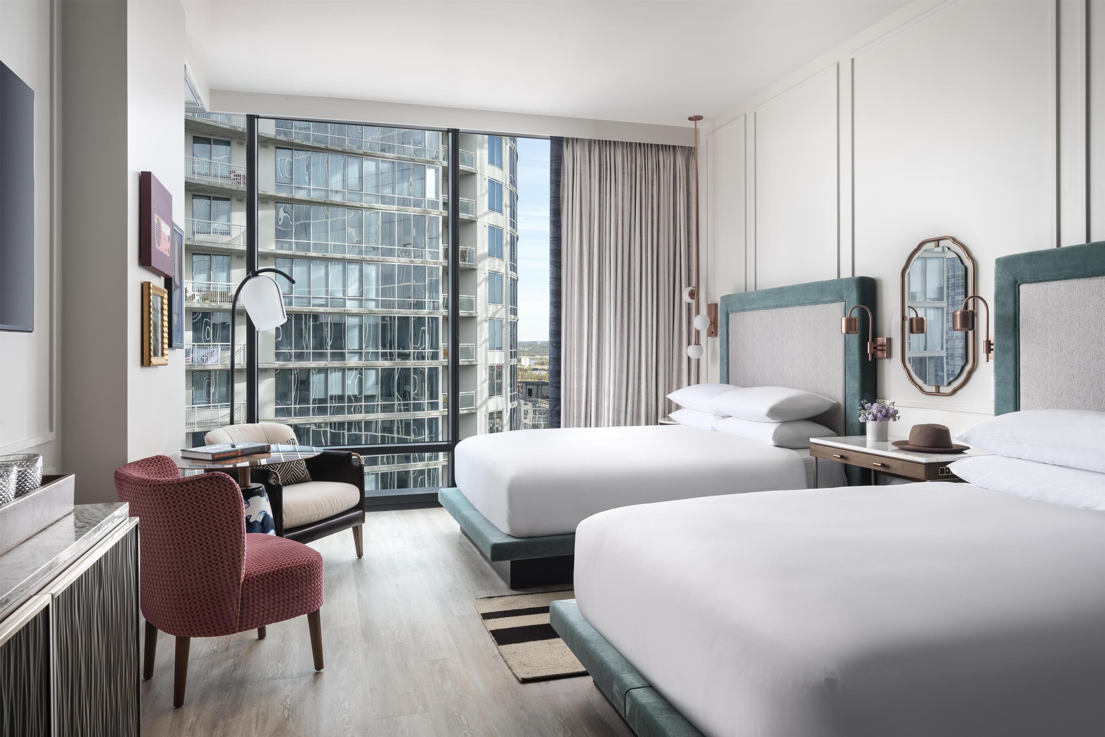 A hotel room with two beds, a desk, a chair, a large window with a view of buildings, and a mirror on the wall.