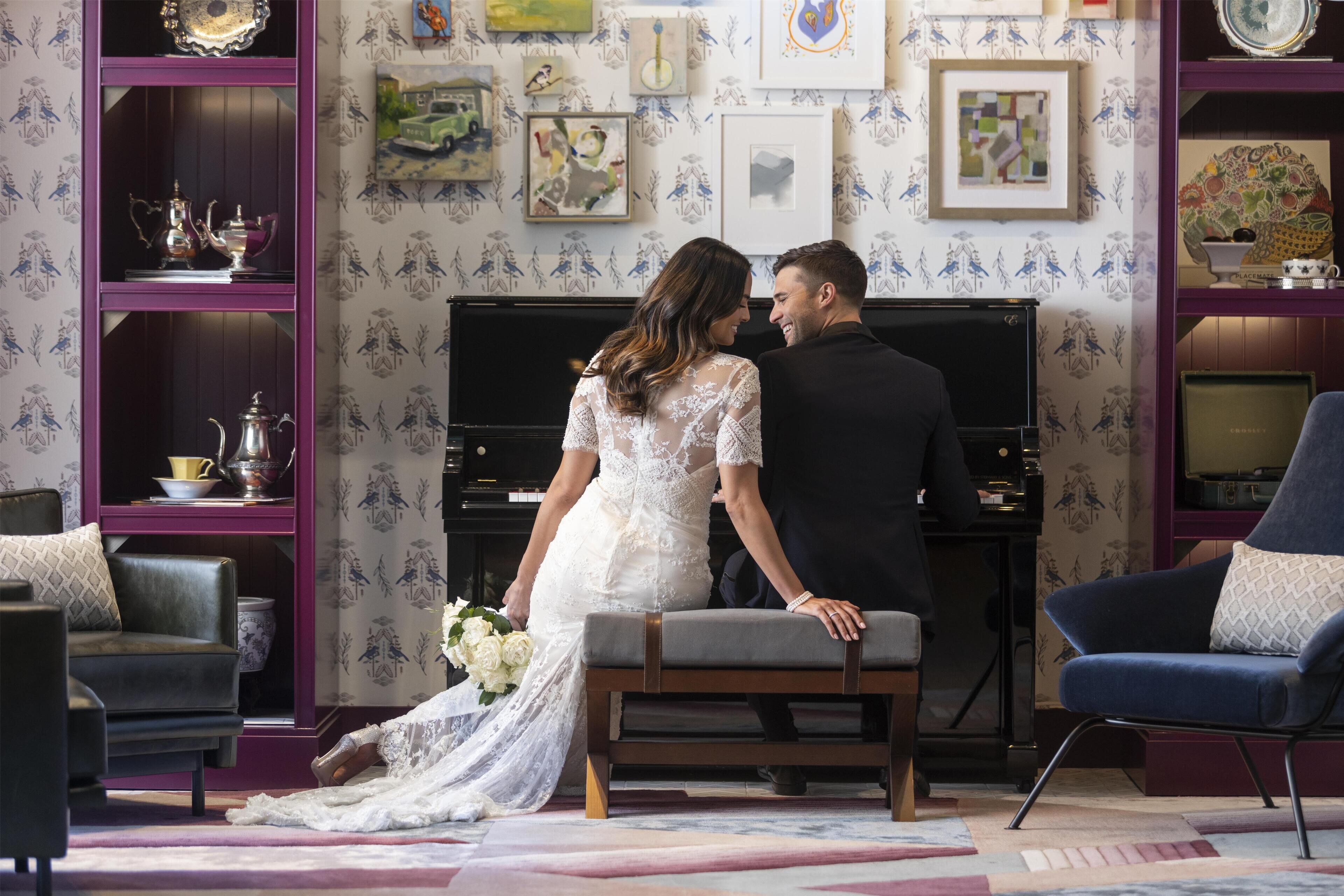 Un couple, vêtu pour un mariage, est assis sur un banc dans une pièce où l’on aperçoit un piano, des œuvres d’art encadrées et un papier peint à motifs.