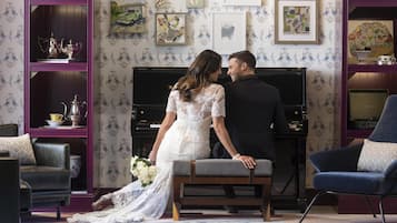 A couple in wedding attire seated on a bench in a room with a piano, framed artwork, and patterned wallpaper.