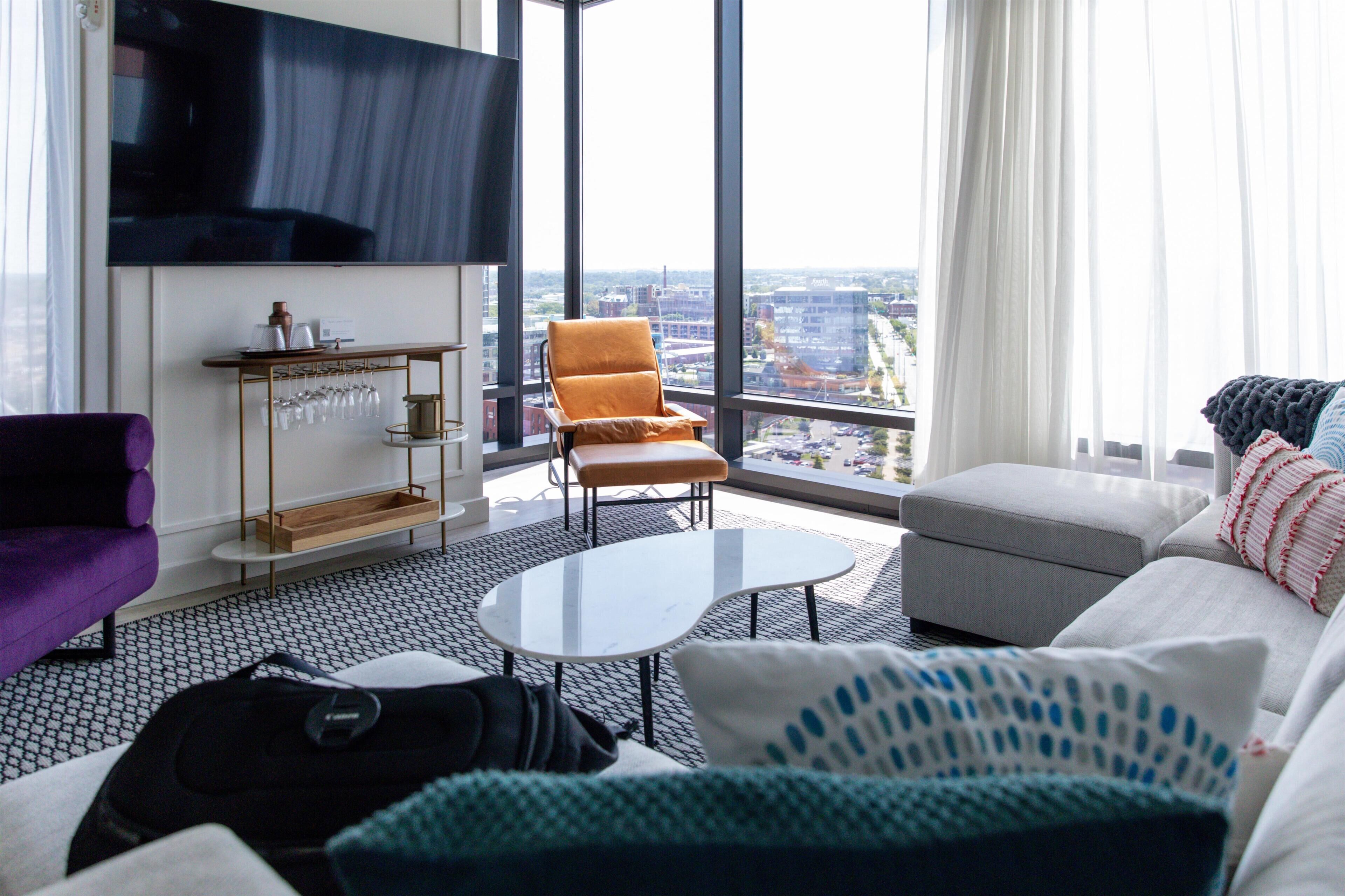 A modern living room with a sofa, armchair, and coffee table, a TV mounted on the wall, and a city view through large windows.