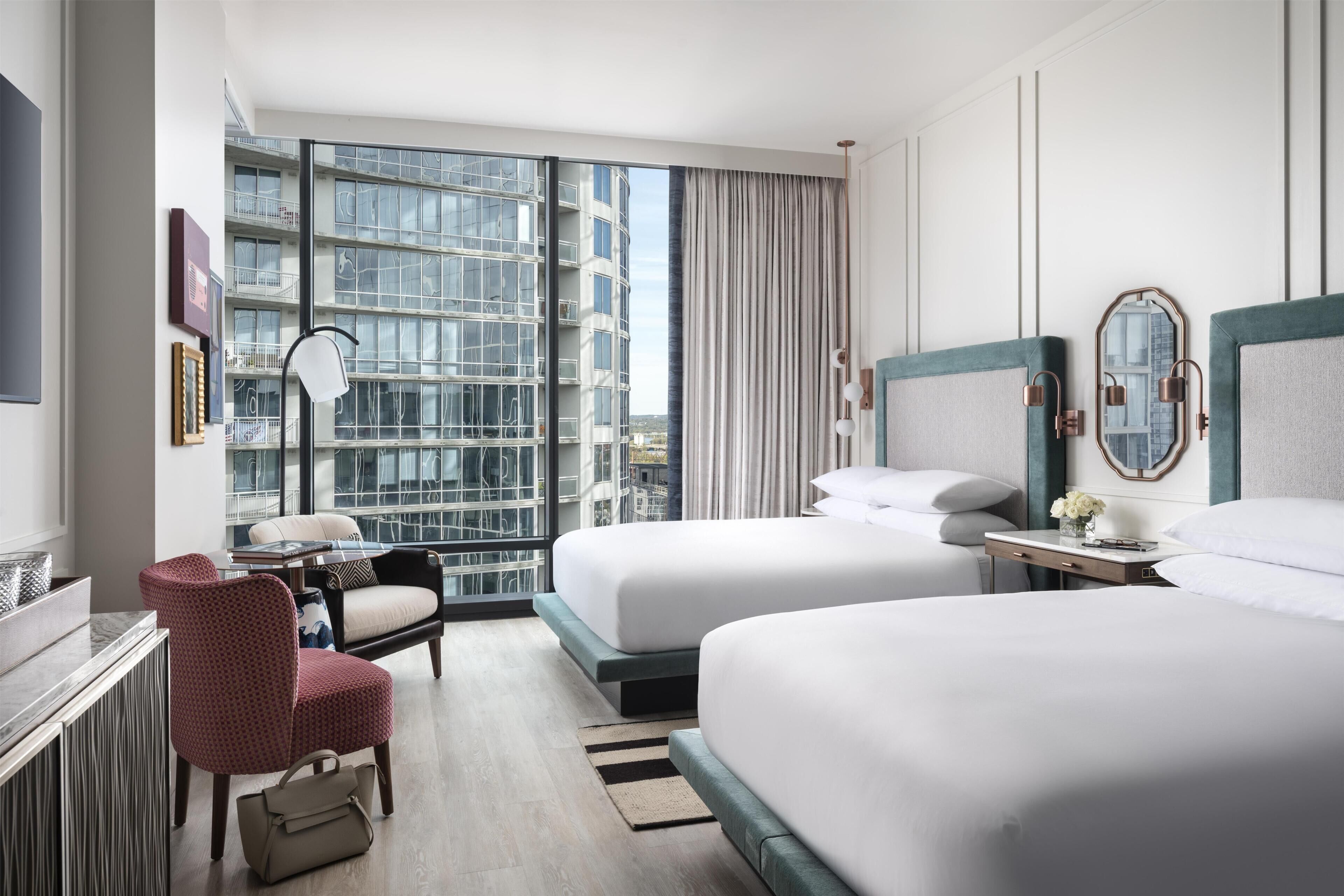 Ett hotellrum med två sängar, ett skrivbord med en stol, ett stort fönster med utsikt över byggnader och en spegel på väggen.