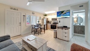 Een moderne keuken met witte kasten, een magnetron en een groot raam. Er is een eethoek met een tafel en stoelen, en een zithoek met een bank en salontafel.