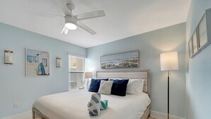 Een net opgemaakt bed met wit- en blauw beddengoed, een plafondventilator en een vloerlamp.
