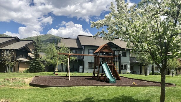 Un terrain de jeux équipé d’un toboggan et de balançoires, entouré d’une pelouse bien entretenue et bordé d’un bâtiment doté d’un toit en pente.