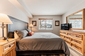 Una camera da letto con un letto, una cassettiera in legno, due lampade da comodino, una televisione e una finestra che si affaccia su alberi e montagne.