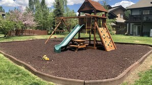 Une aire de jeux avec un toboggan et des balançoires, entourée d’une zone recouverte de paillis et d’une clôture en bois.