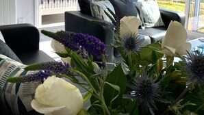 A living room with a sofa, a coffee table with a floral arrangement, and a view of the outside through large windows. - 2 Bed Villa Sleeps 4 - Wifi, Garden, Sea Views (Looe)