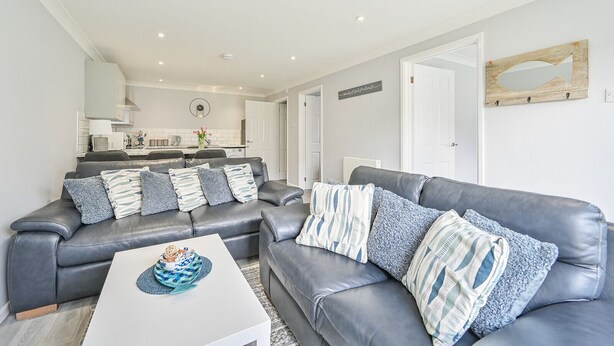 A modern living room with a grey leather sofa, a white coffee table, and a kitchen in the background. - 2 Bed Villa Sleeps 4 - Wifi, Garden, Sea Views (Looe)
