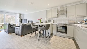 A modern kitchen with white cabinets, a built-in oven, and a dining area with a table and chairs. - 2 Bed Villa Sleeps 4 - Wifi, Garden, Sea Views (Looe)