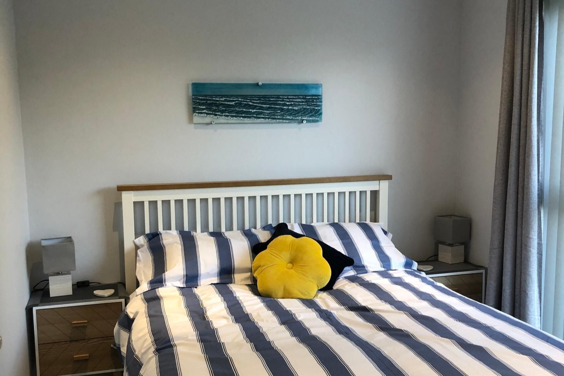 A bed with striped bedding, a yellow pillow, a white headboard, a bedside table with a lamp, and a wall-mounted artwork.