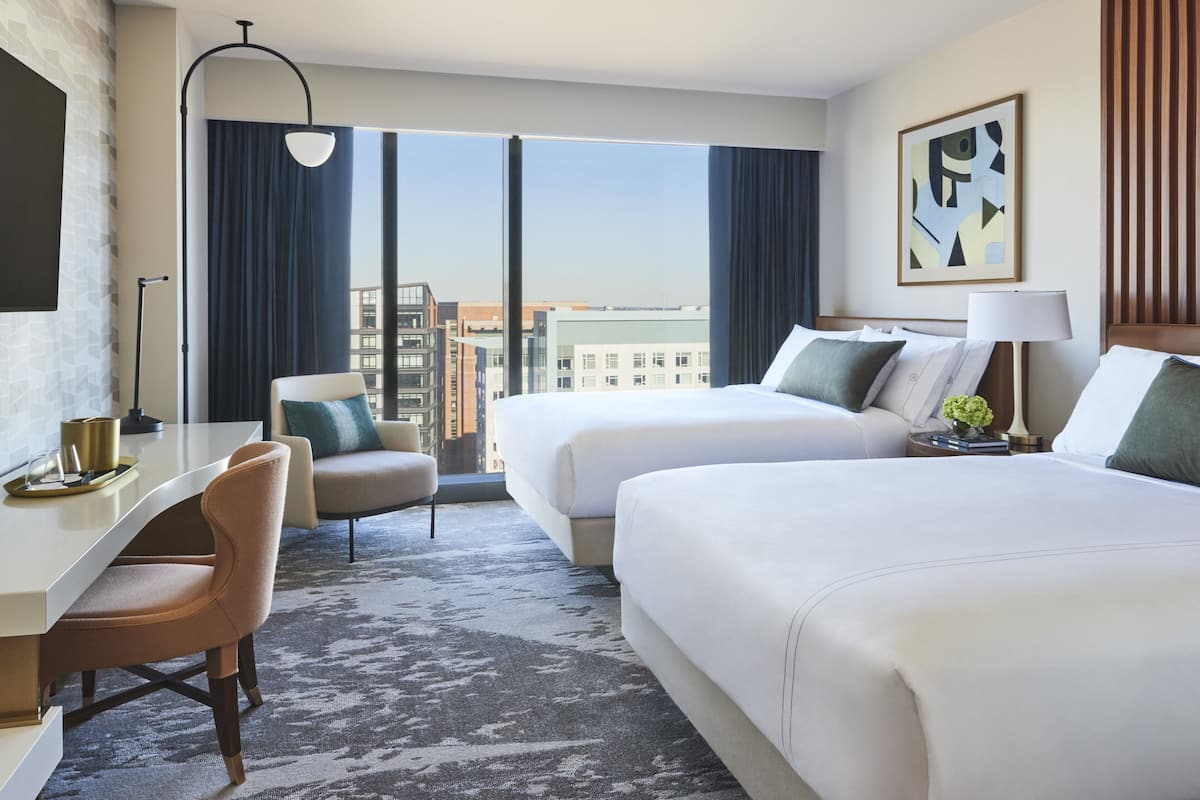 A hotel room with two beds, a desk, a chair, and a large window with a city view.