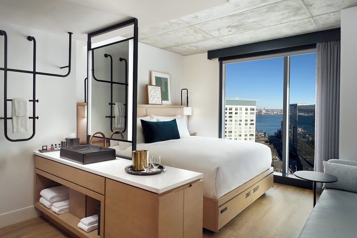 A modern hotel room with a large bed, a vanity with a mirror, a small table, and a view of the city and water.