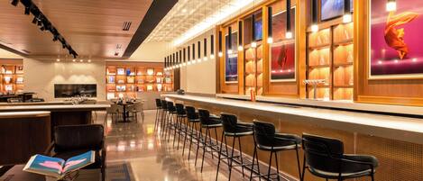 A modern bar area with a long counter, multiple bar stools, and a variety of glassware displayed in wooden cabinets.
