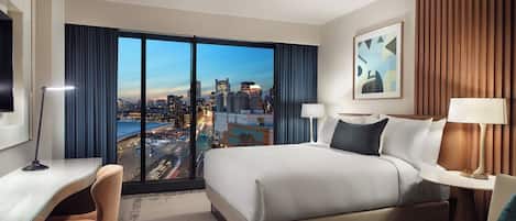 A hotel room with a large bed, a desk, a chair, and a view of a cityscape through a large window.