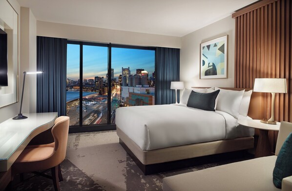 Une chambre d’hôtel avec un grand lit, un bureau, une chaise et une vue sur le paysage urbain, visible à travers une grande fenêtre.
