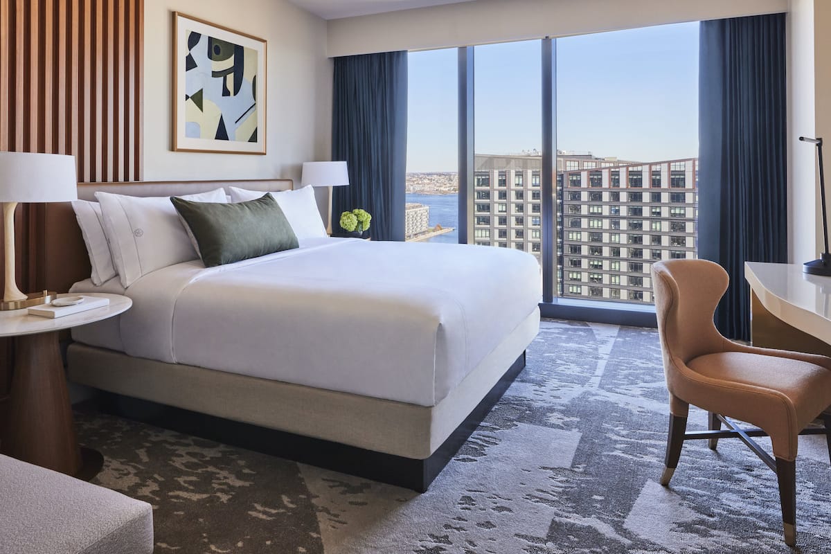 A hotel room with a large bed, a desk, a chair, and a view of a cityscape through large windows.