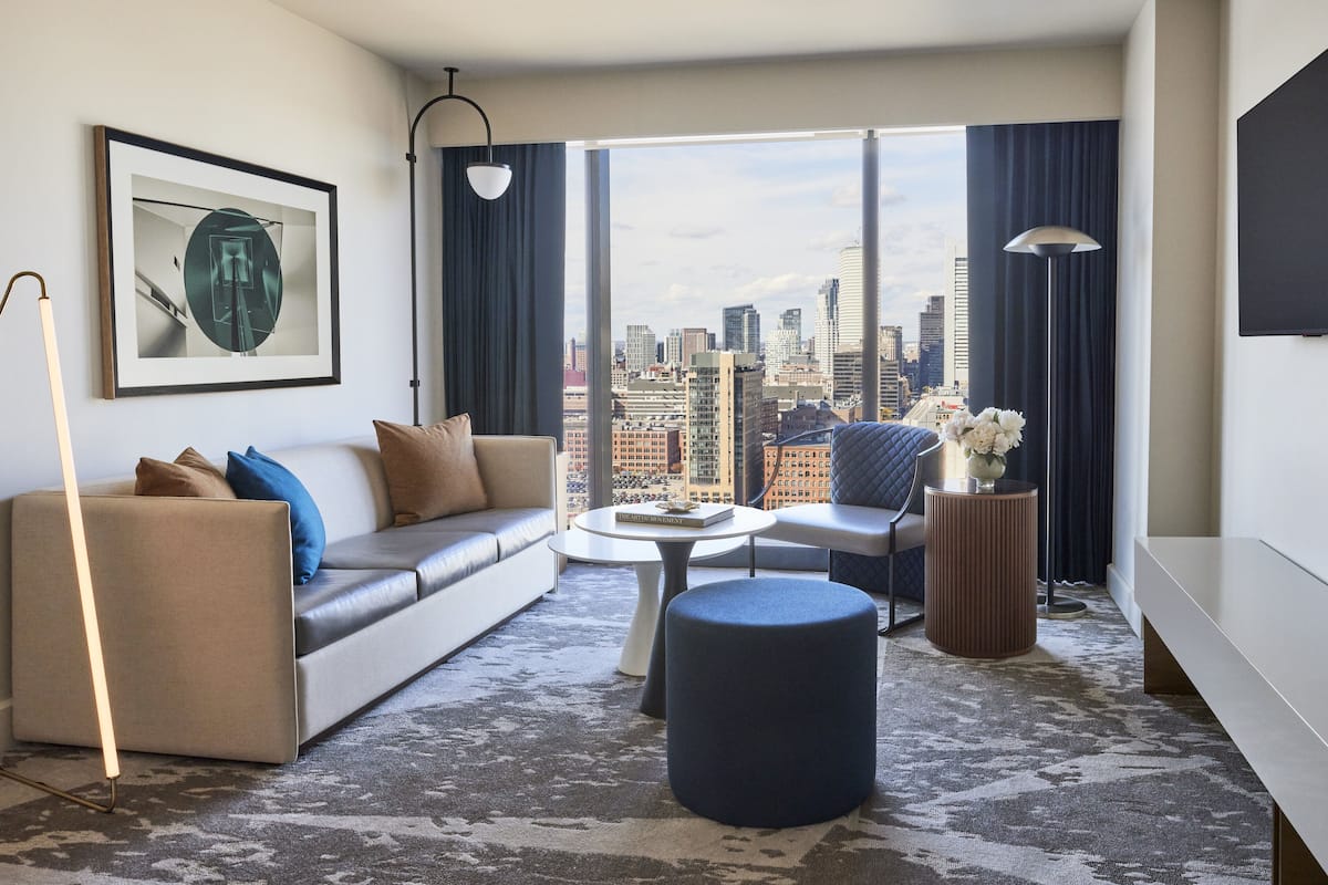 A modern living room with a sofa, ottoman, and a coffee table. A large window offers a city view.