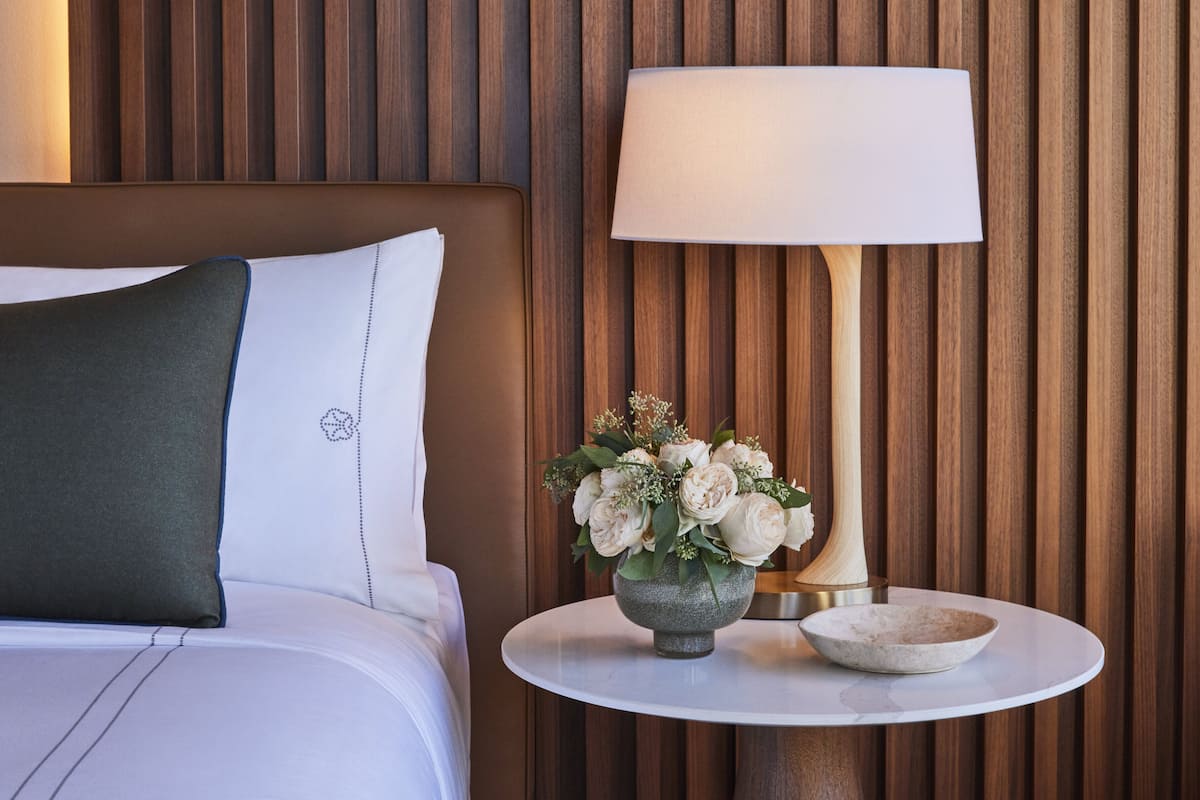 A neatly made bed with white linens and a dark green pillow, a bedside table with a lamp and a vase of flowers, and a wooden panel wall.