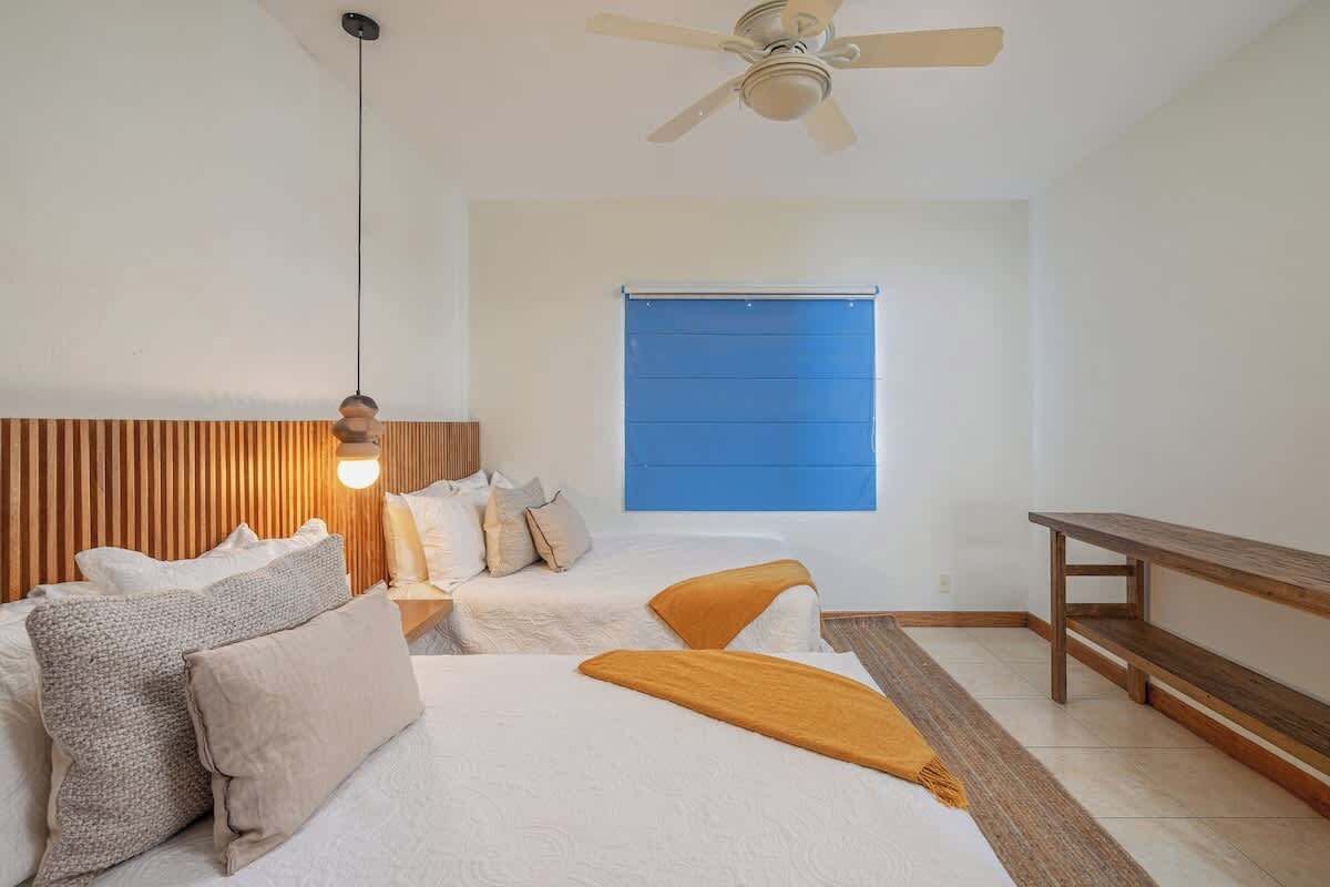 A bedroom with two beds, a ceiling fan, a window with blinds, and a wooden bench.