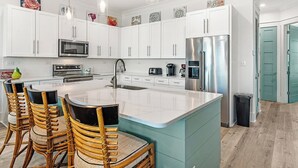 A modern kitchen with white cabinets, stainless steel appliances, a central island, and a dining area with wicker chairs. - 😎 Night Heron, West. ⭐️⭐️⭐️⭐️⭐️ 6BR. Heated Pool. Beachview. Gorgeous! (Gulf Shores)