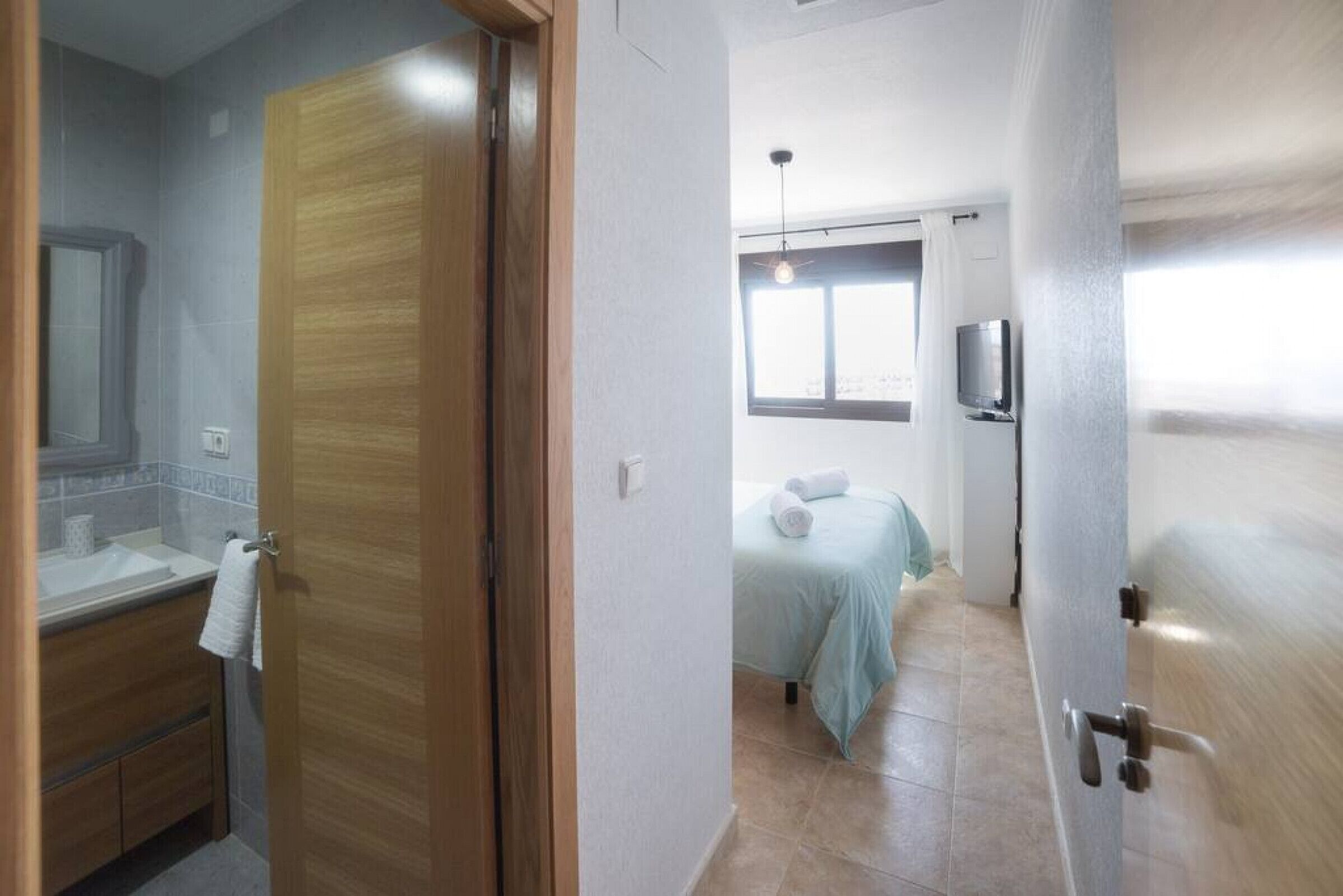 Ein Hotelzimmer mit Holztür, einem Badezimmer inklusive Waschbecken und Spiegel, einem Bett mit hellblau gestreckter Bettdecke sowie einem an der Wand befestigten Fernseher.