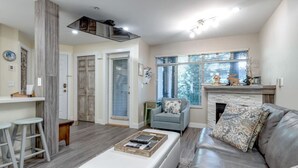 A modern living room with a grey leather sofa, a white ottoman, and a blue armchair. There is a fireplace with decorative items and a window with blinds.