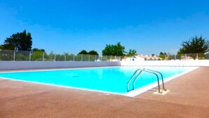 Une piscine rectangulaire avec un escalier en métal sur un côté, entourée d’une zone délimitée par une clôture, avec des arbres et des bâtiments en arrière-plan.