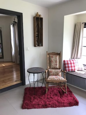 Una habitación con una silla de madera, una mesita redonda pequeña, un tapete rojo, un radiador y una ventana con cortinas.