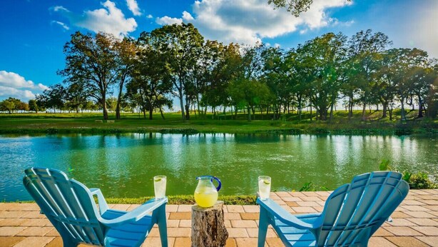 Two blue chairs facing a pond with a pitcher of lemonade between them, surrounded by trees and a clear sky.