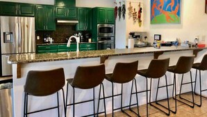 A modern kitchen with green cabinets, stainless steel appliances, a granite countertop, and a bar area with stools.