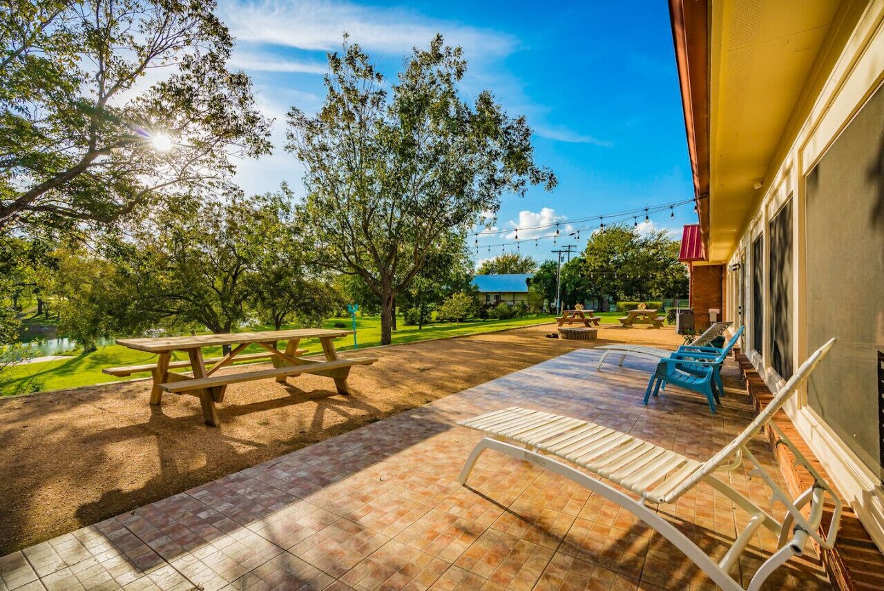 An outdoor patio with a picnic table, lounge chairs, and a view of a park with trees and a pond.