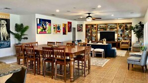 A spacious living room with a wooden dining table, comfortable seating, a television, and a bookshelf filled with books and decorative items.