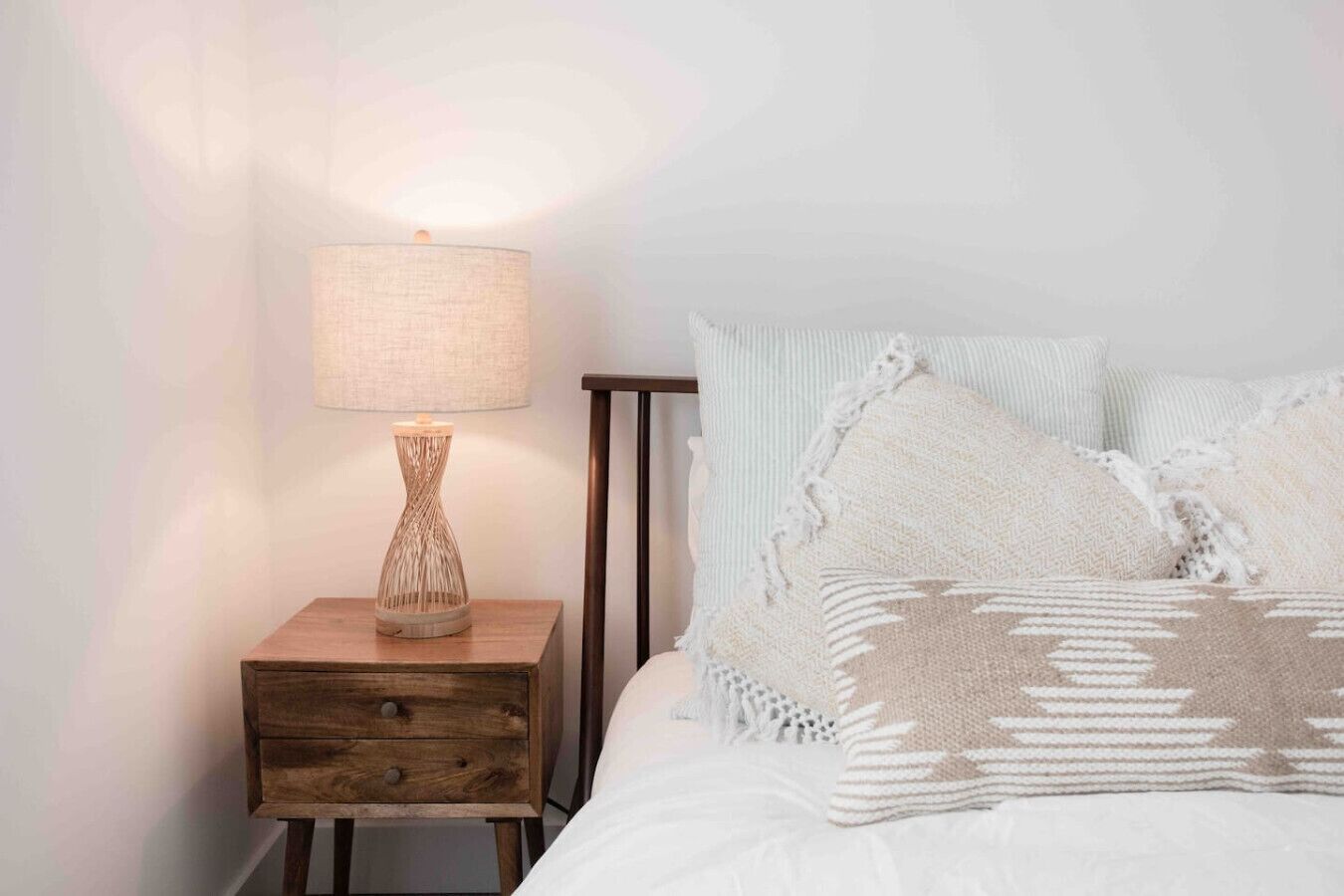 A neatly made bed with a white comforter and a patterned throw blanket. A wooden nightstand with a lamp and a vase is beside the bed.