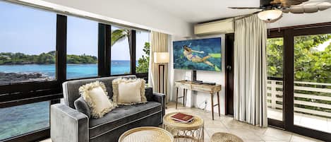 A modern living room with a large window overlooking a scenic ocean view, a ceiling fan, and a flat-screen TV.