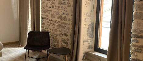 A room with a stone wall, a bed, a chair, and a window with curtains.