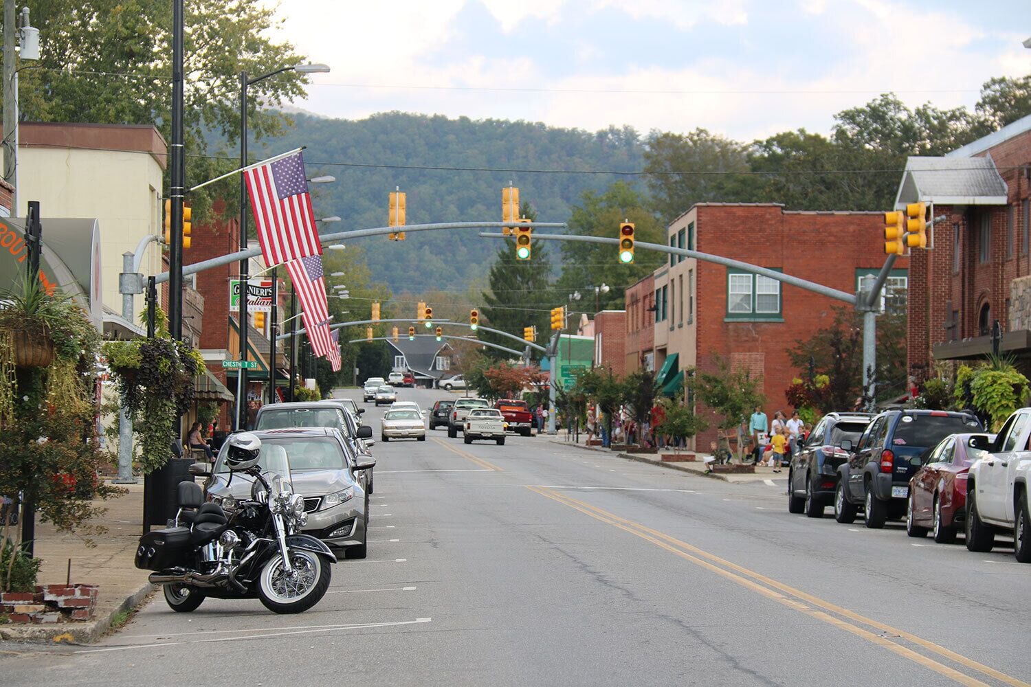 A street with cars parked on the side, a motorcycle, and an American flag. There are buildings on both sides of the street, and a traffic light is visible.