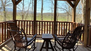 Eine Holzterrasse mit zwei Stühlen und einem Tisch, mit Blick auf eine ländliche Landschaft mit Bäumen und Hügeln.