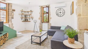 Un salon avec un canapé gris, une table basse en bois et un téléviseur posé sur un meuble. On y trouve des étagères ornées d’objets décoratifs et une grande horloge au mur.